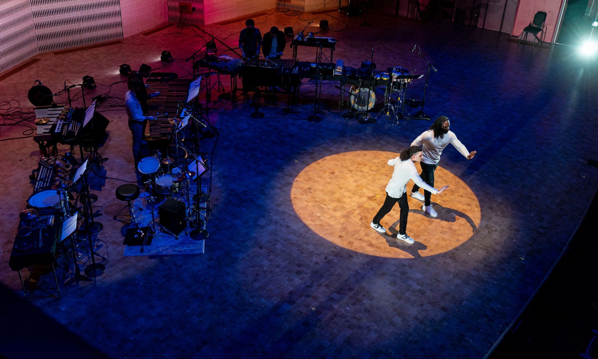 Two dancer dressed in white long sleeved shirts and black skinny jeans with white sneakers raise one arm forward, facing the audience