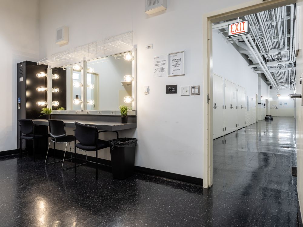 Image of Dressing Room 2. There is a vanity mirror station with chairs and a black locker to the left of the vanity. Through the open door on the right-side you can see the hallway. 