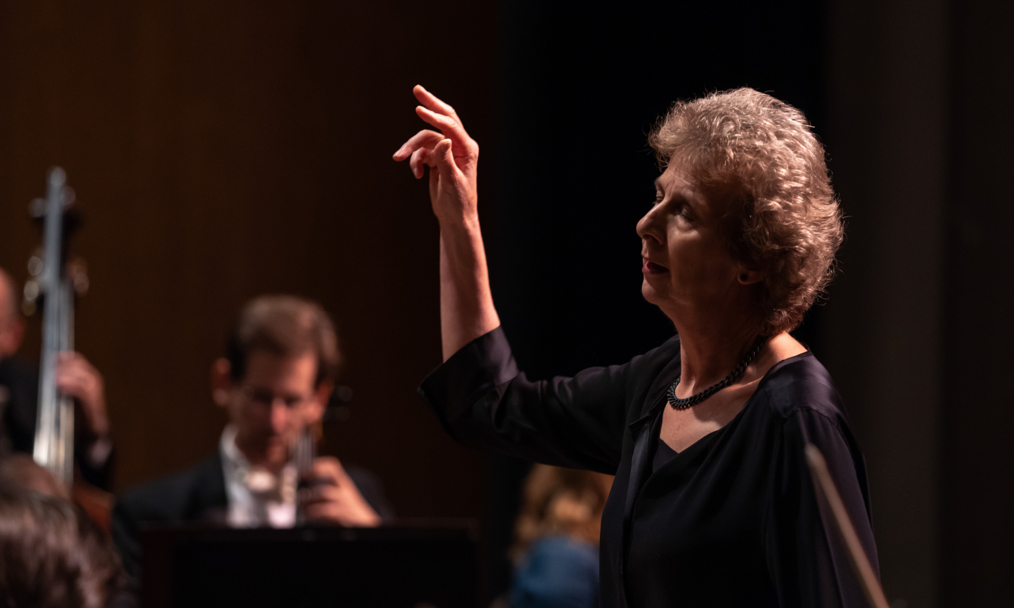 Dame Jane Glover stands with her eyes closed and right arm raised, mid-conducting.