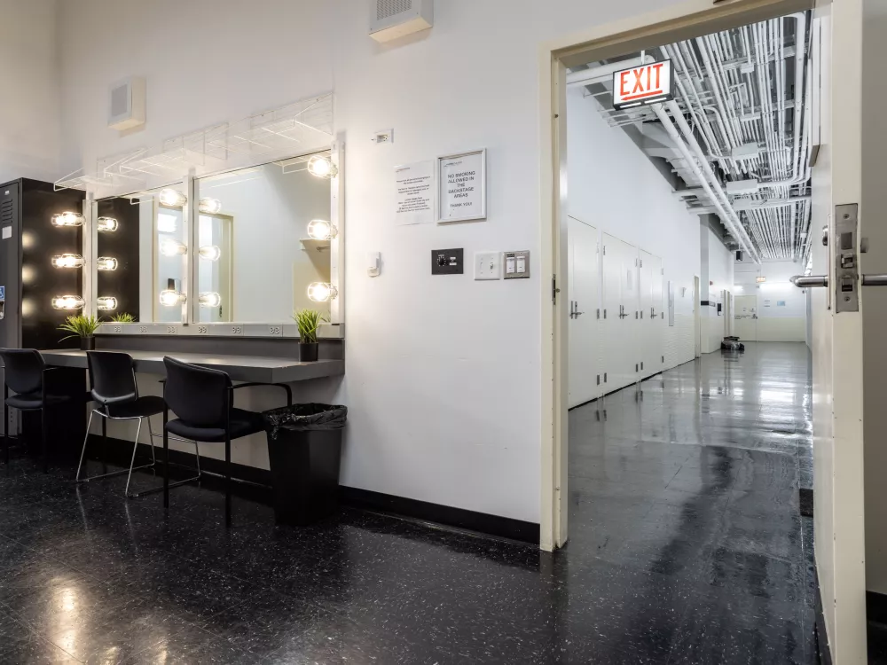 Image of Dressing Room 2. There is a vanity mirror station with chairs and a black locker to the left of the vanity. Through the open door on the right-side you can see the hallway. 