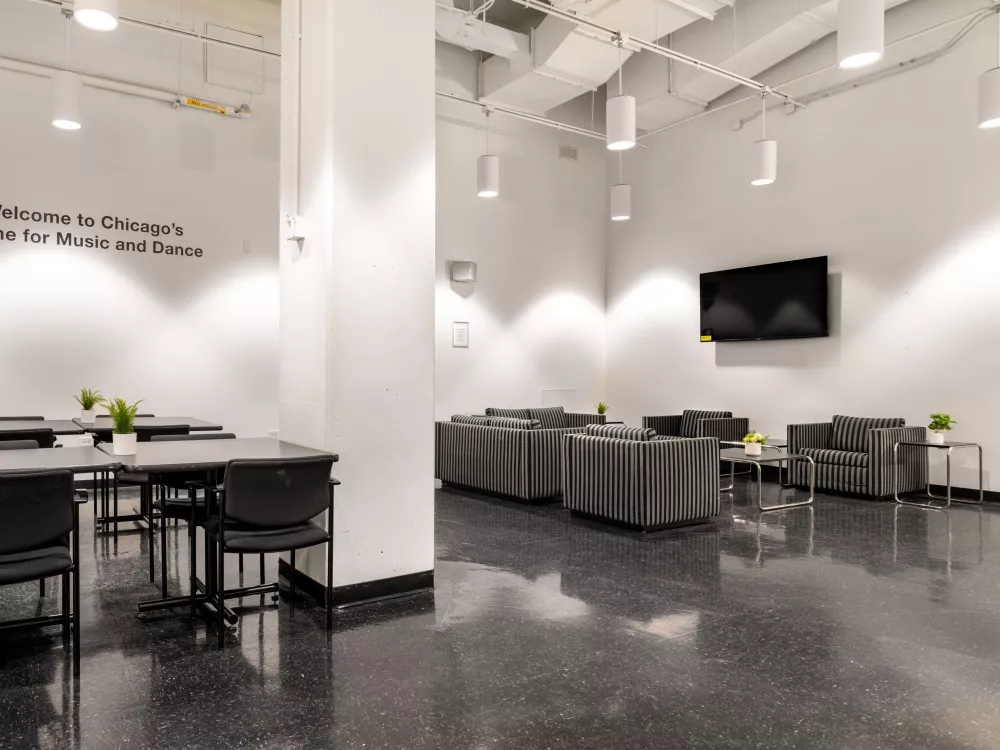 Image of an empty lounge room. On the left-side of the room, four tables with chairs are placed. On the right-side of the room four stripped couch chairs with side tables are placed. A tv hangs the wall of the right-side of the room.