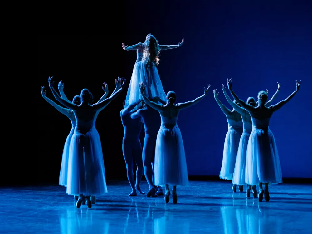 A group of ballerinas raise their arms. One dancer is lifted and also raising her arms while leaning her head back.