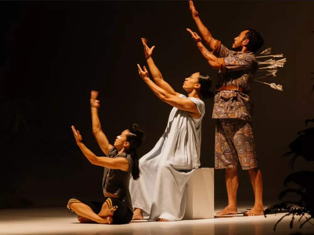Three dancers extend their arms up to the right in unison. One sits on the floor, another on a box right behind the first dancer, and the third stands in the back of all three.
