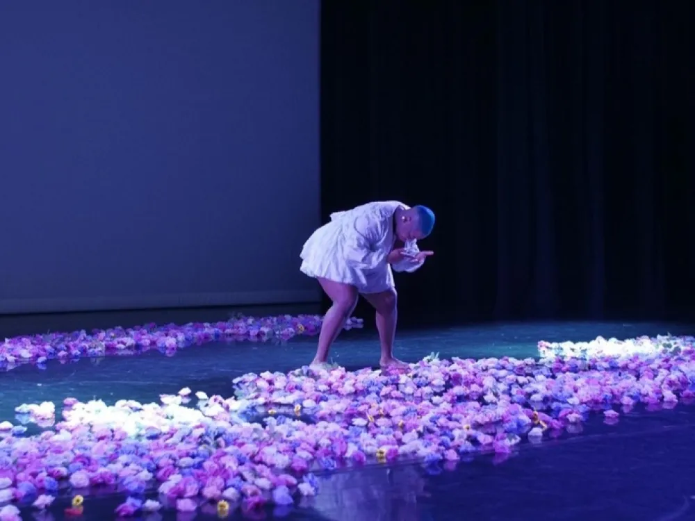 A performer on stage bending and looking down. There are flowers all over the floor.