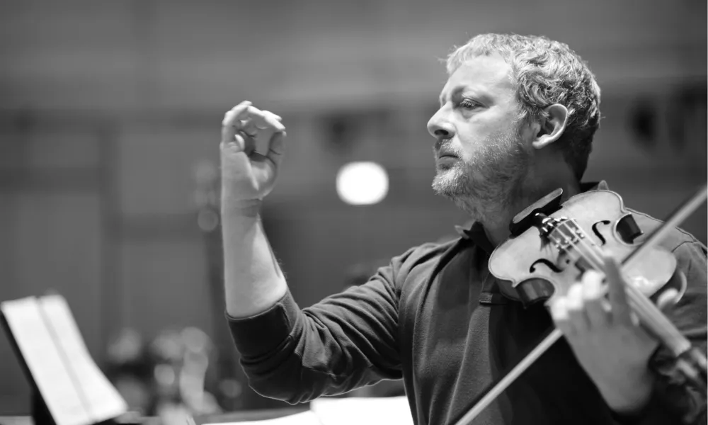 Black and white photo of Fabio Biondi holding his violin with his other hand raised.