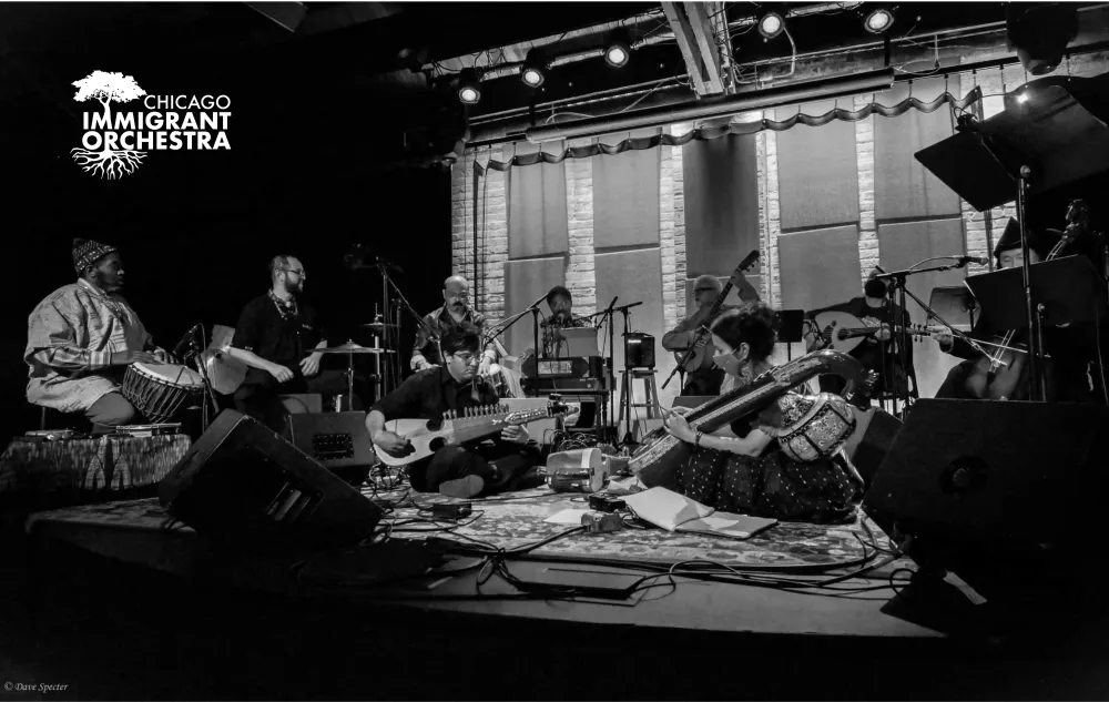 A black-and-white photo of the Chicago Immigrant Orchestra showing six musicians sitting in a semi-circle and a stage playing various string and percussion instruments.