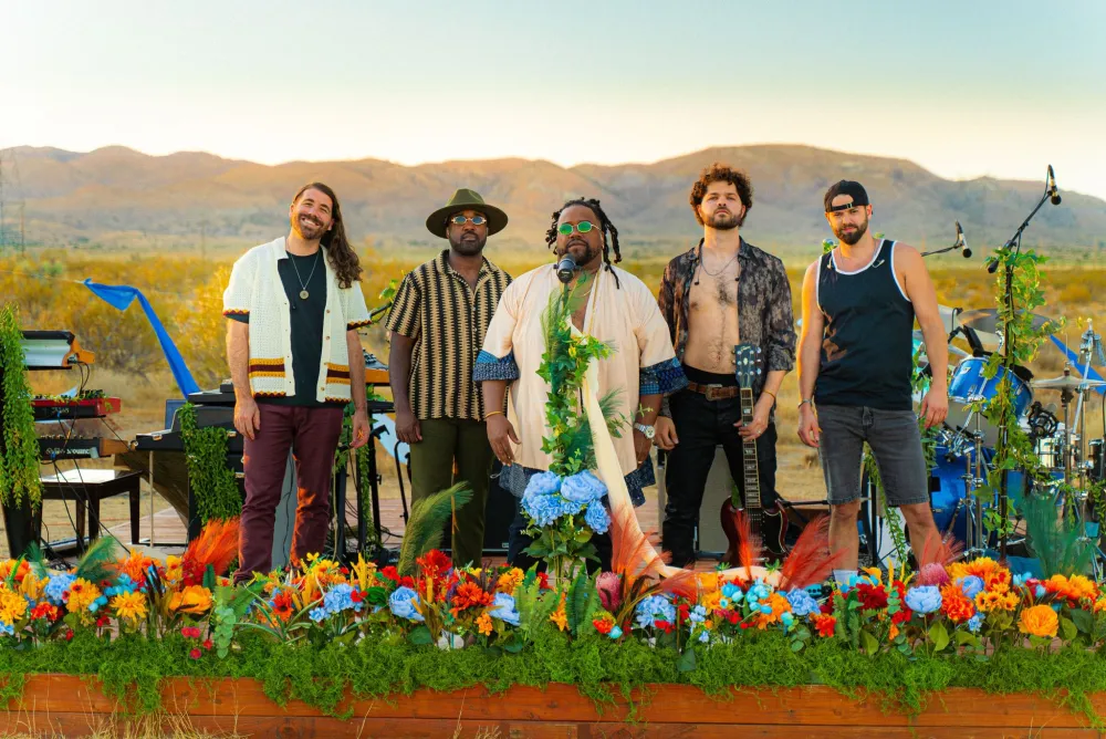 Five men on a stage in the dessert. The stage is filled with greenery, flowers, and their instruments..