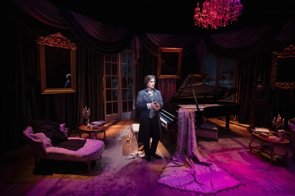 A man in a dark suit stands in a dark room that looks like a salon with a large piano. 