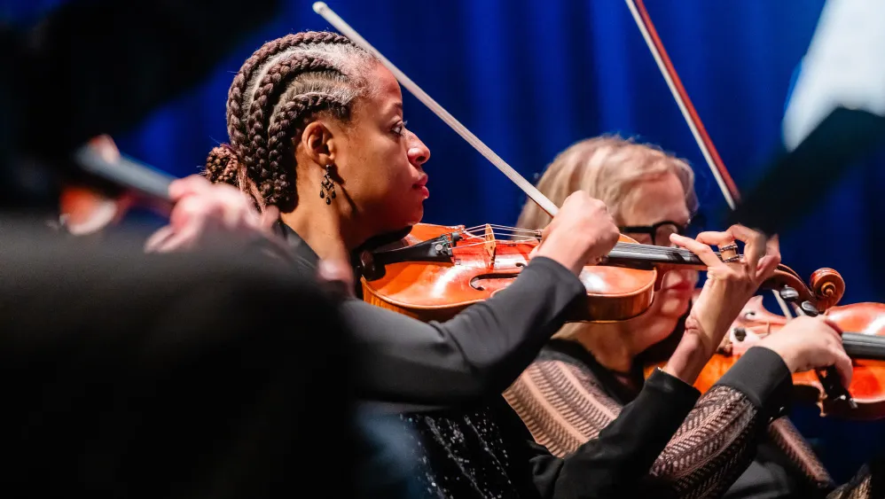 Violinists perform on stage, focused on their instruments as bows move in unison under blue stage lighting.