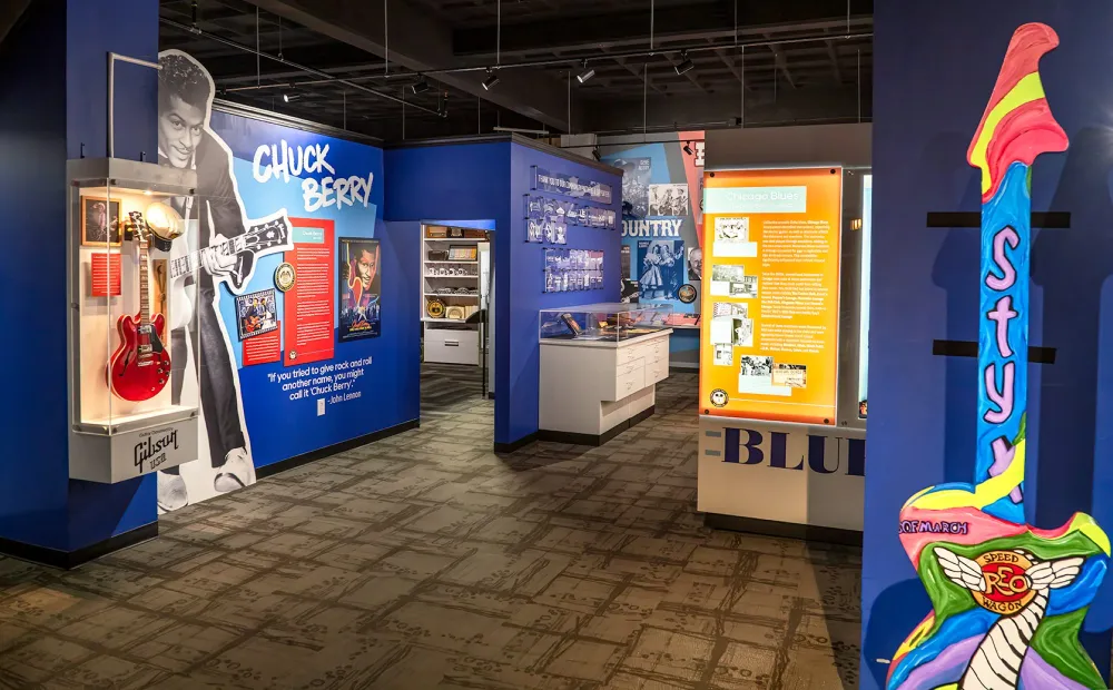 A museum exhibit features a large Chuck Berry display with a life‑size photo of him performing, a red Gibson guitar in a glass case, and framed photos and text panels. Nearby sections highlight Chicago blues and country music, and a colorful oversized guitar sculpture painted with STYX and REO Speedwagon imagery stands to the right.