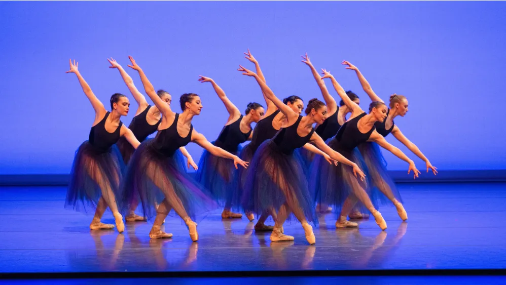 A large ensemble of ballet dancers in flowing dark blue costumes moves in unison across a stage bathed in blue light. With arms extended diagonally upward and bodies leaning forward, the dancers create a sweeping sense of momentum, precision, and collective energy.
