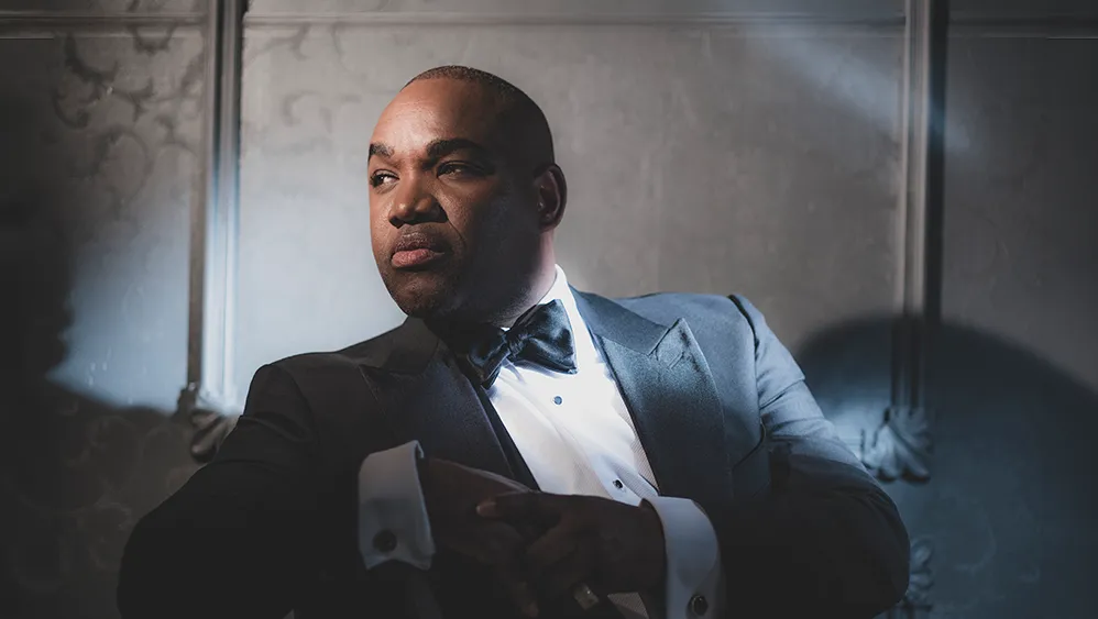 A man sitting in a tuxedo sitting down and looking to his right. He rest his arm on the top of the chair.
