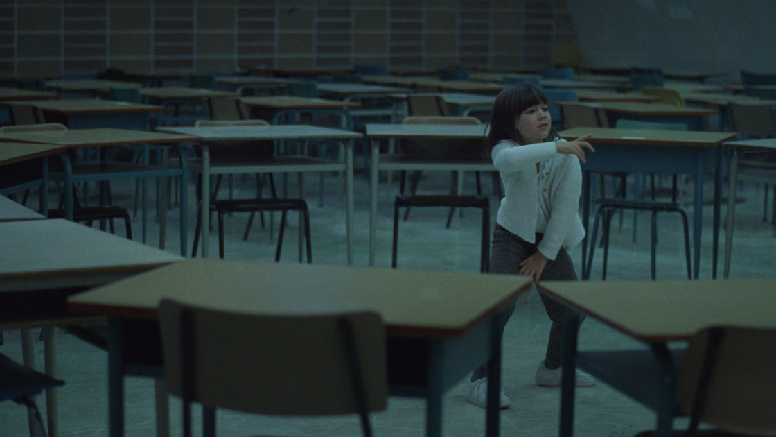 A young girl with dark hair and a white sweater dances, surrounded by desks in a classroom