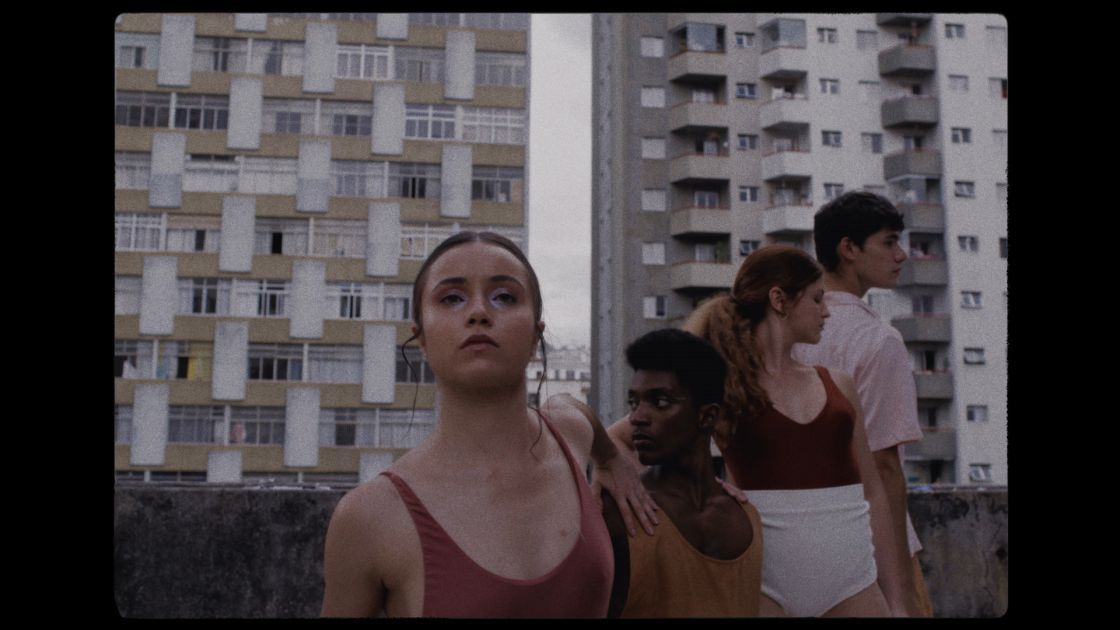 Four dancers stand close together in a diagonal row on a rooftop, tall buildings flanked behind them in a gray sky. Female dancers wear solid red or gold tops, and one male dancer wears a cream button down. One dancer looks at the camera while the others look  to the sides. 
