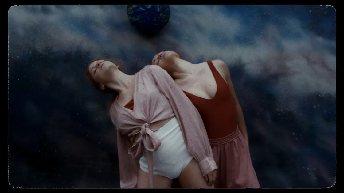 Two dancers stand side-by-side close together, one dancer slightly behind the other. Both dancers are leaning far left with eyes closed, focused upwards. Dancers wear shades of deep red, pink, and white and stand in front of a swirling painted background of dark blue, light blue, and white. A blue orb is seen in the painting above the dancers' heads.