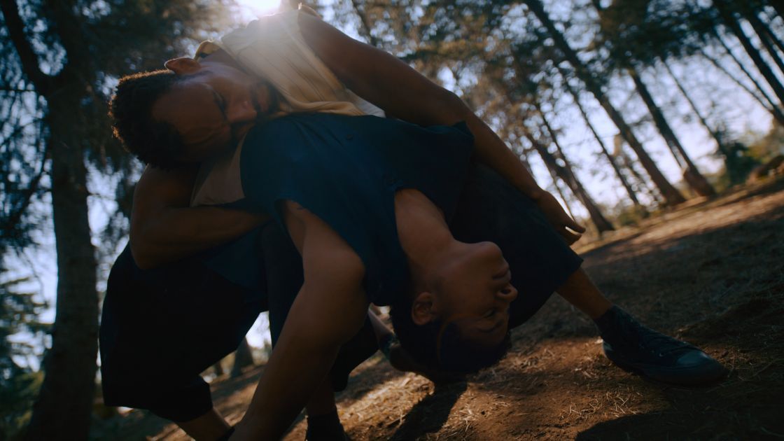 A Black man in a white shirt and a White woman in a blue shirt dance in a wooded setting at dusk