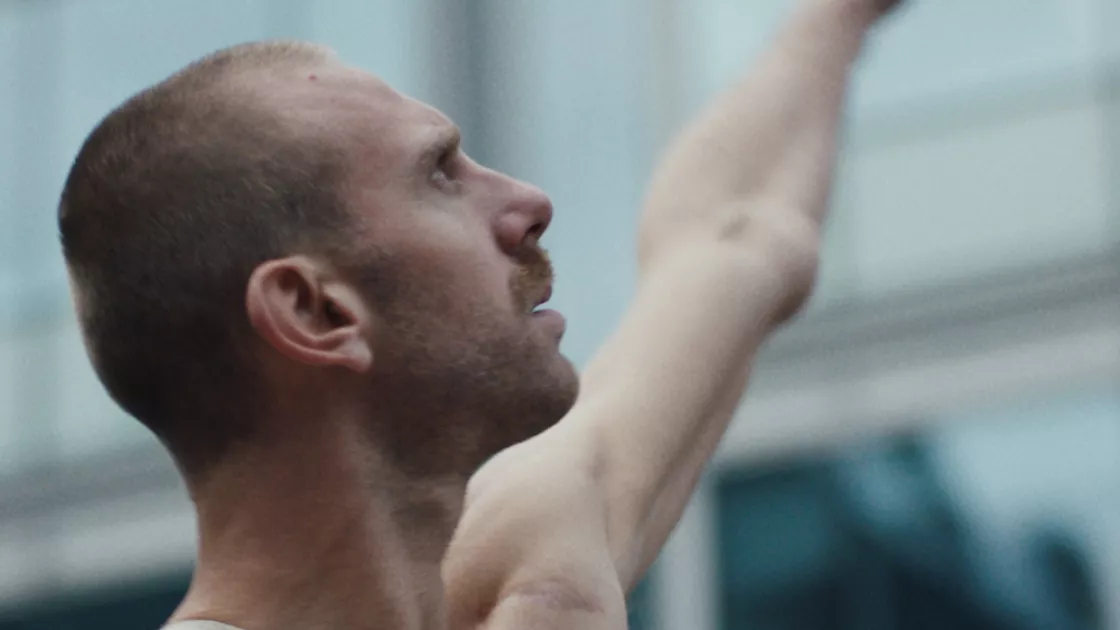 One dancer stands in profile in front of a glass building, their left arm extended diagonally out to their side. Their right arm with limb loss extends straight out from their body. Dancer's eyes are focused upwards.