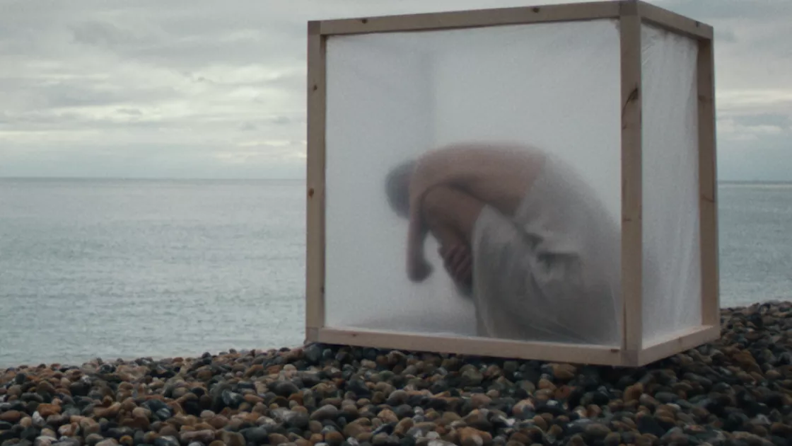 A single dancer sits inside a translucent square box, with arms wrapped tight around legs, head tucked down. Box is positioned on a rocky shore, a cloud-filled sky and gray body of water can be seen in the background.