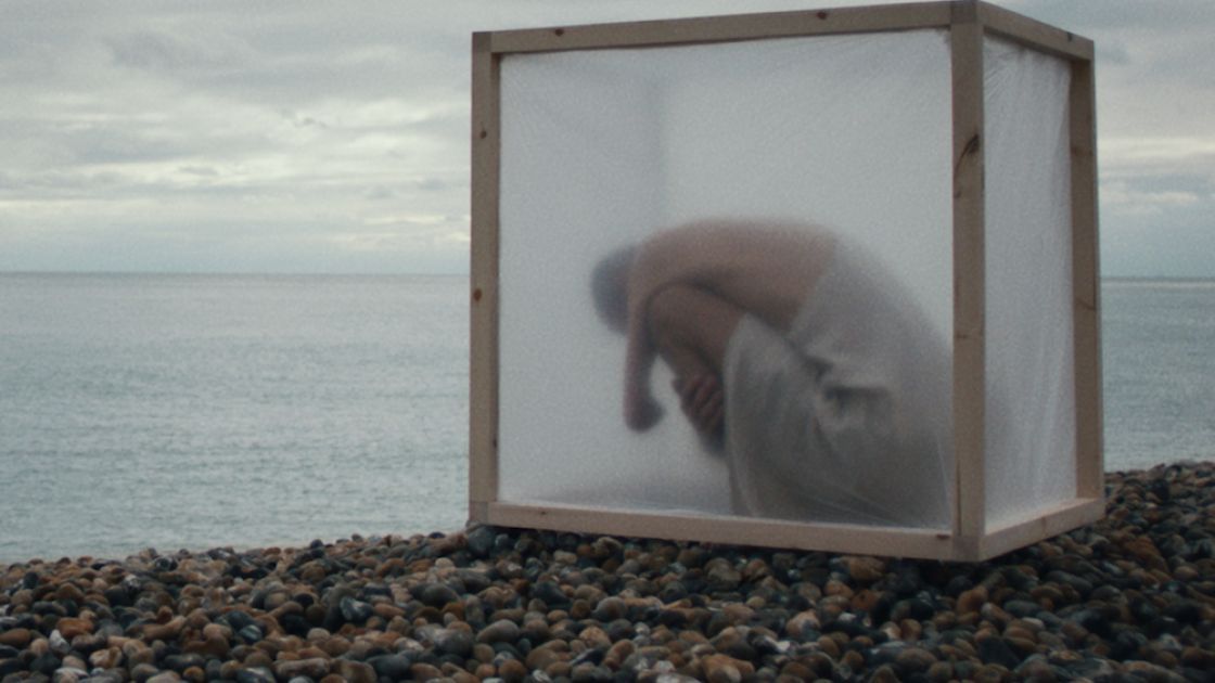 A single dancer sits inside a translucent square box,  with arms wrapped tight around legs,  head tucked down.  Box is positioned on a rocky shore, a cloud-filled sky and gray body of water can be seen in the background.