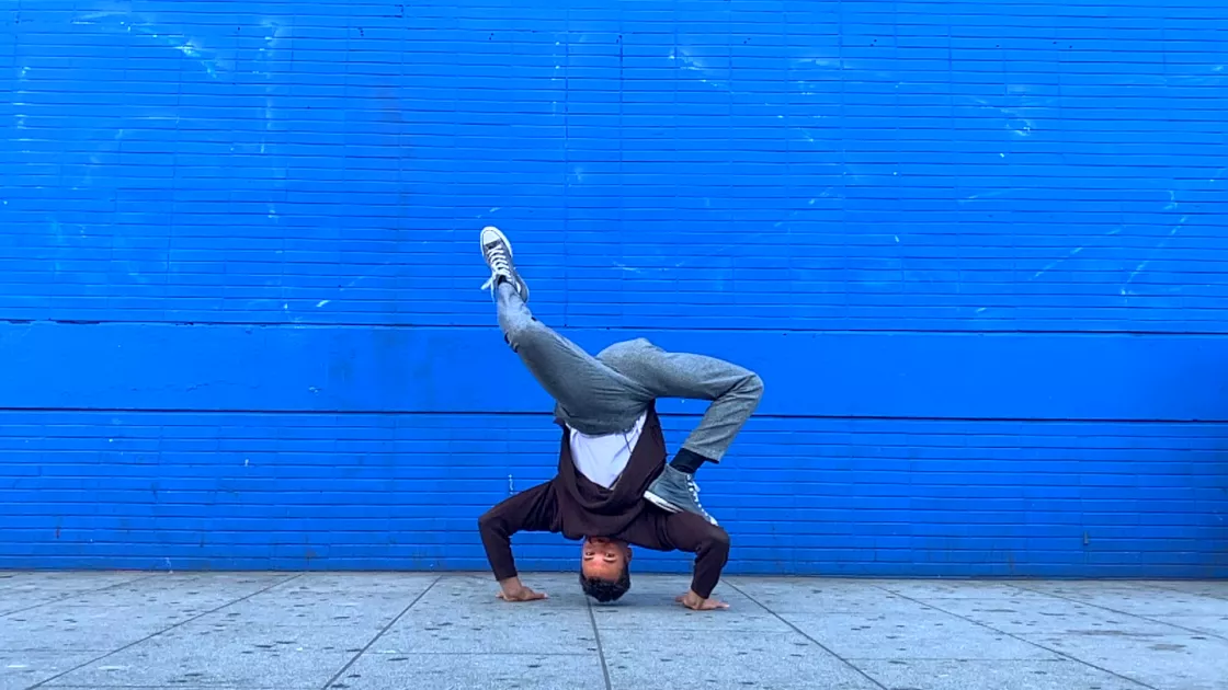 A dancer in jeans and a dark shirt does a headstand on concrete, against the backdrop of a bright blue wall