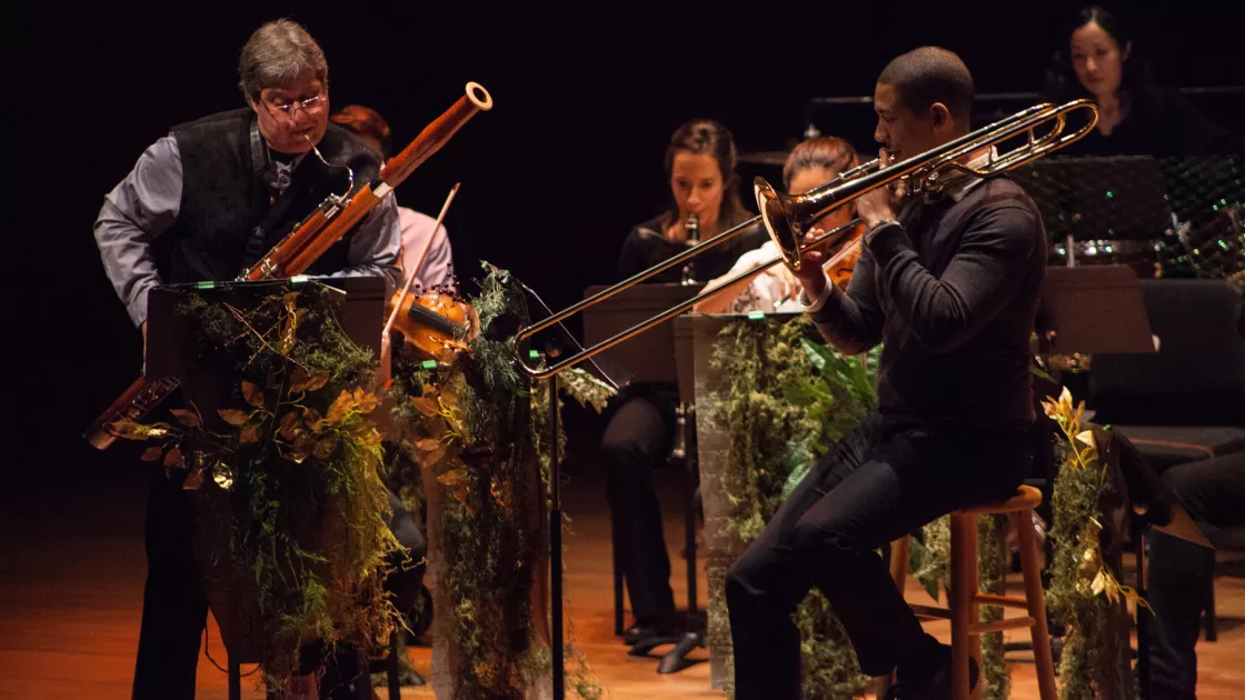 An oboe player and a trombone player perform in the foreground with other musicians in the background.