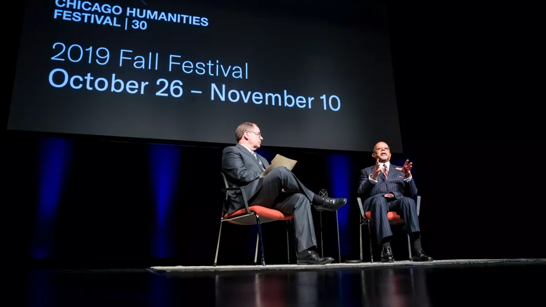 Two men, one White and one Black, sit in chairs on a dark stage, against a backdrop that reads Chicago Humanities Festival - 2019 Fall Festival