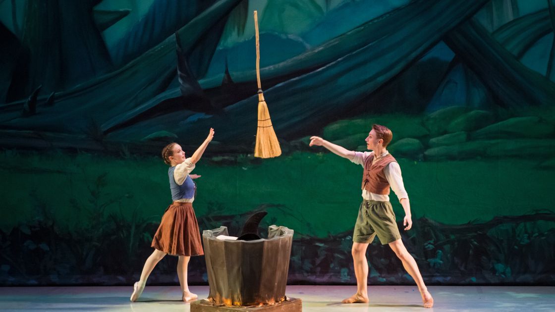 One male and one female dancer stand facing each other against a green, woods-like backdrop, one tossing a broom to the other