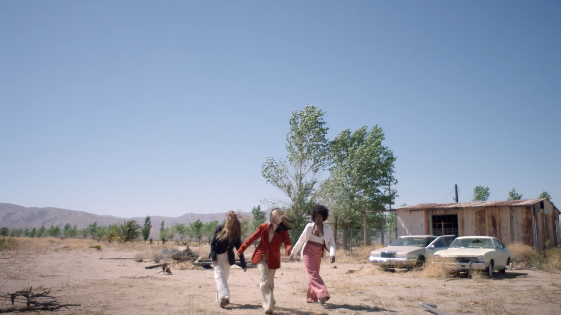 Three dancers moving their right foot forward in a desert-like town with two cars and a house