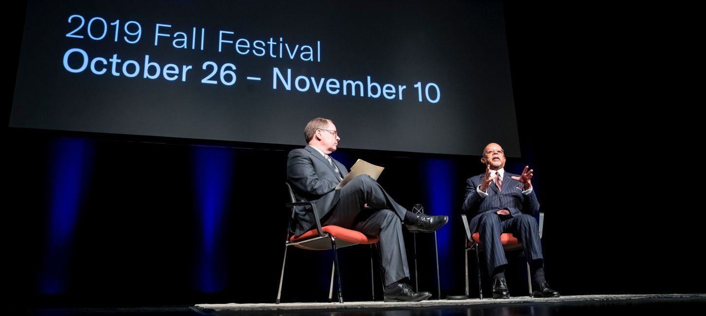 Two men, one White and one Black, sit in chairs on a dark stage, against a backdrop that reads Chicago Humanities Festival - 2019 Fall Festival