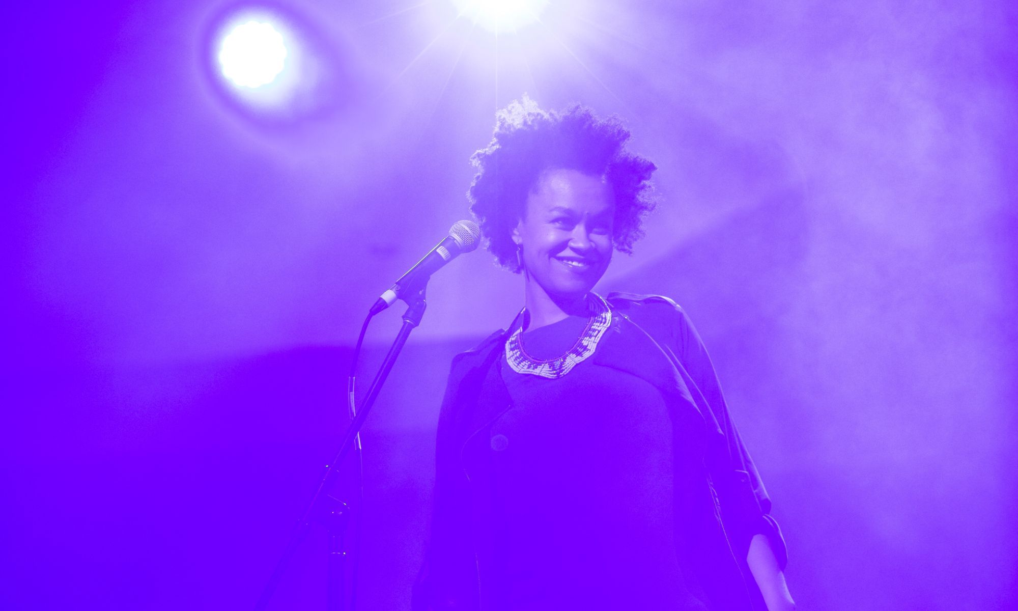 Meklit smiling with a microphone in front of her surrounded by purple lighting