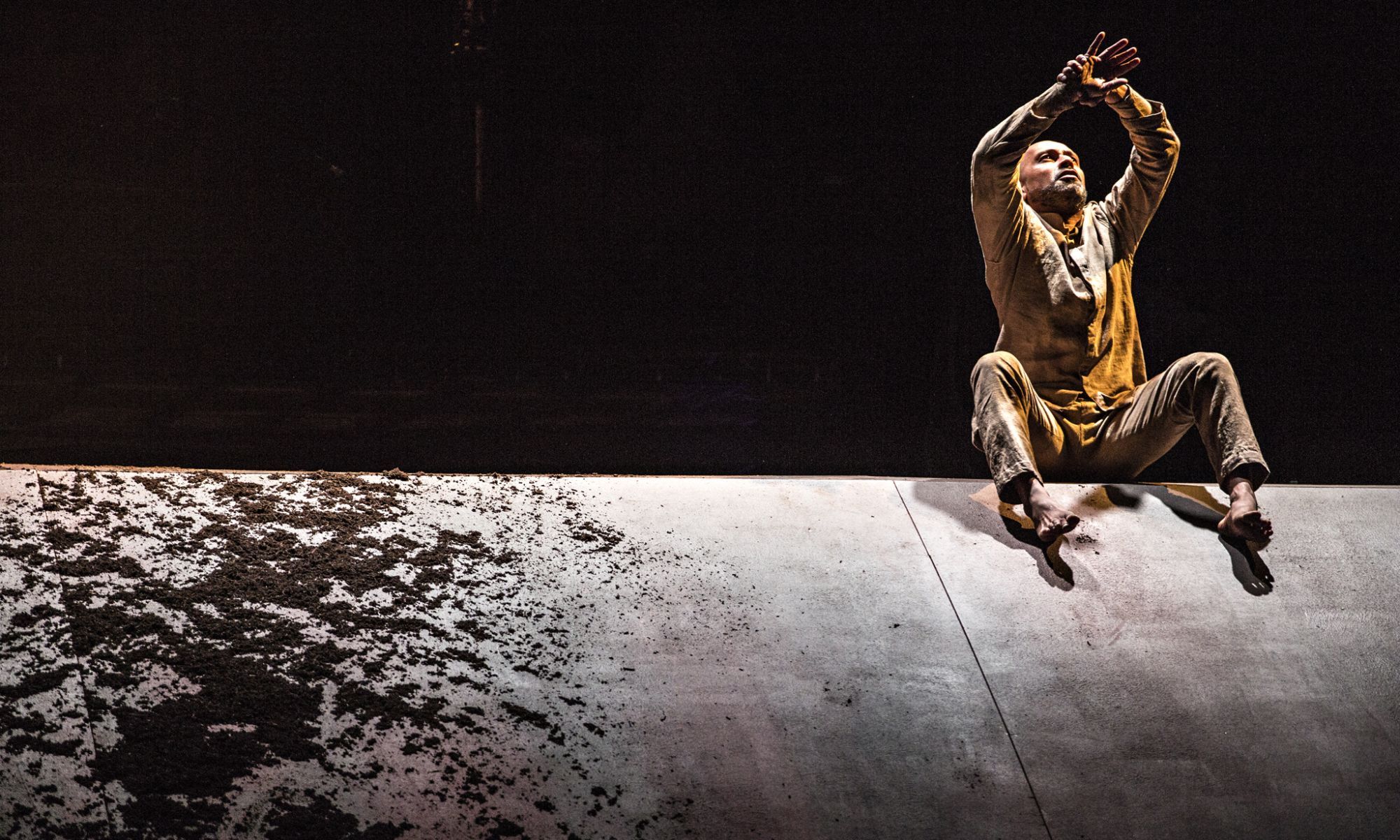 Akram Khan sits on top of a grey wall grasping his wrists overhead