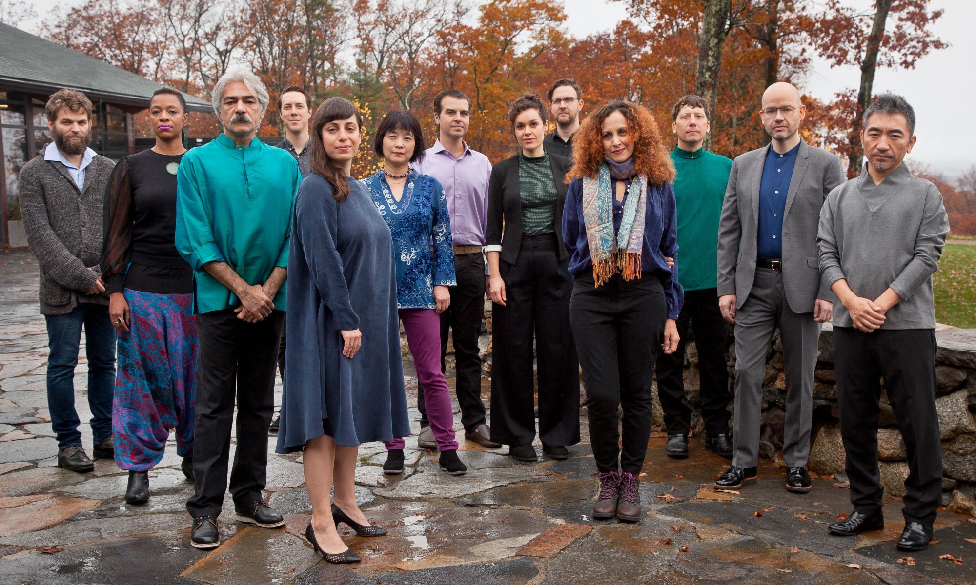 13 musicians in casual attire pose in front of trees with autumn colors