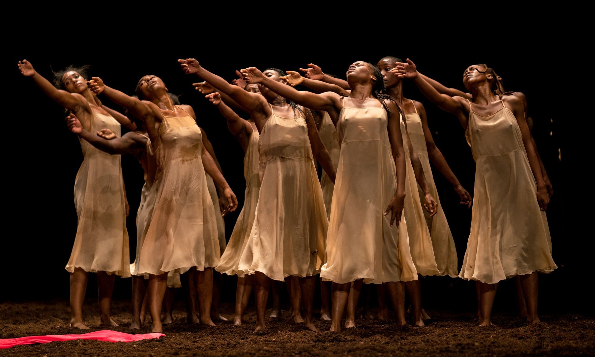Eight Black women dancers in light dresses dance on a dark stage