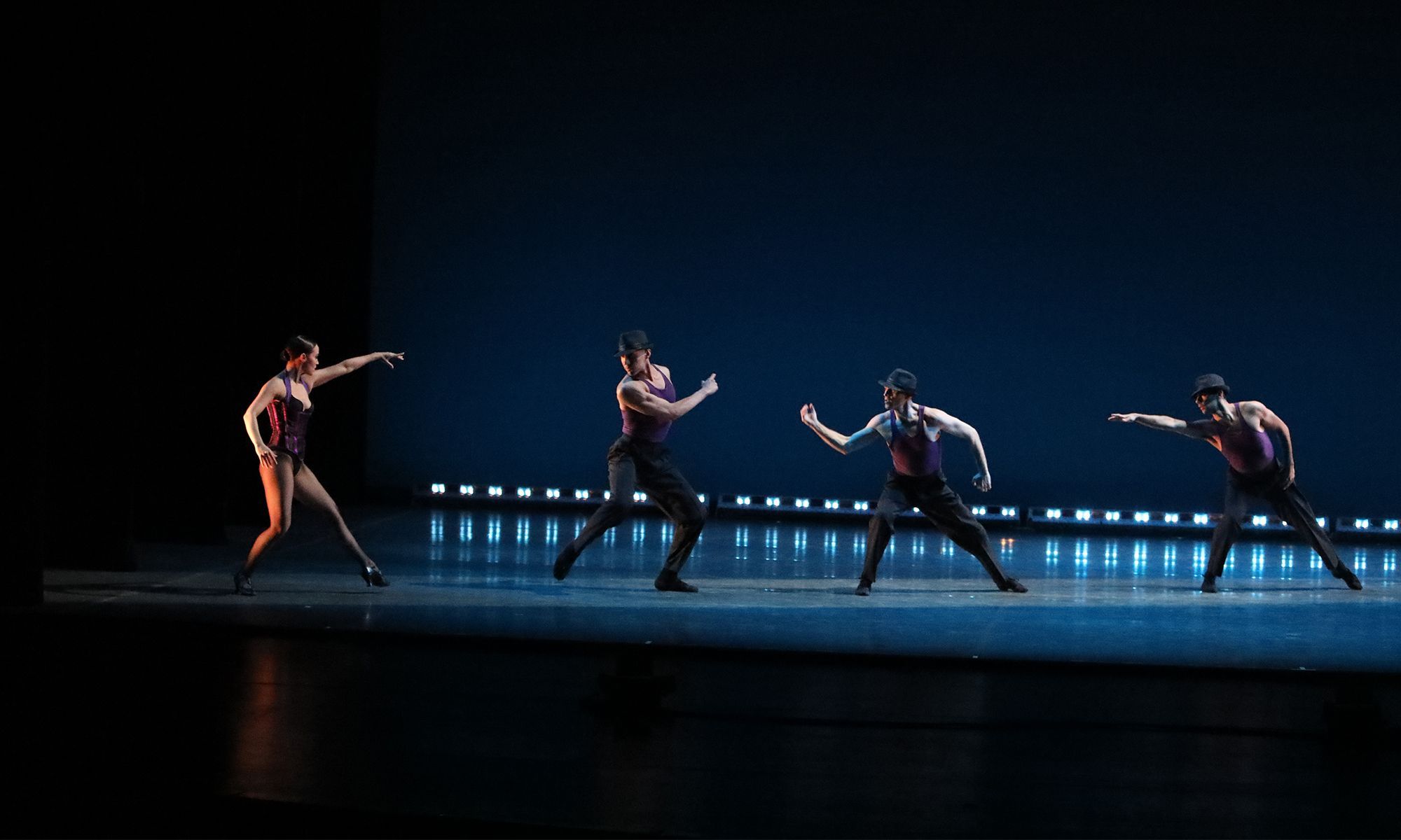 Female dancer in a corset and fishnet tights reaches toward a diagonal of male dancers dressed in pants, tank tops and fedora hats. The stage picture is complete with footlights across the back of the stage. 