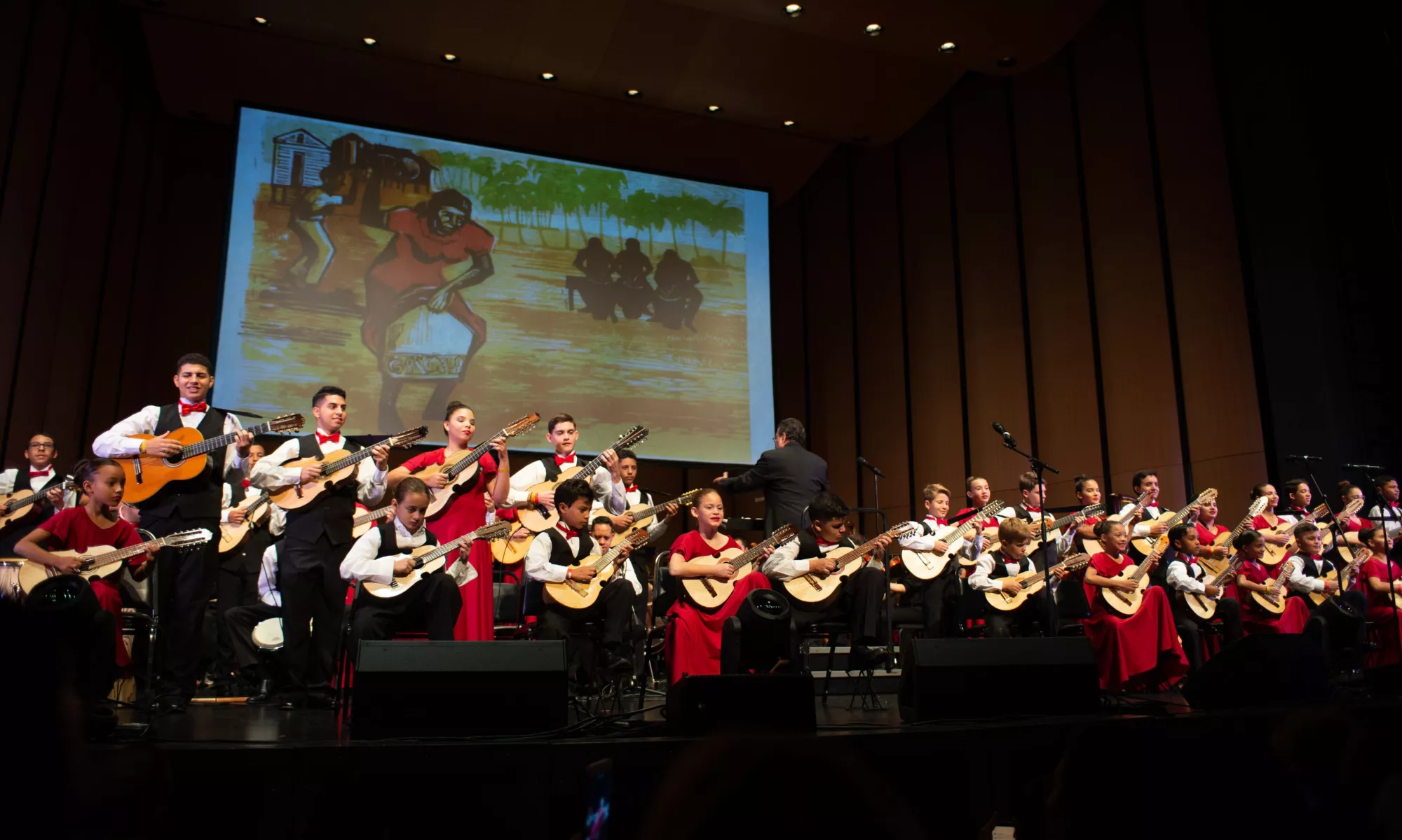 Girls wearing red dresses and boys wearing white shirts with black vest suits playing cuatro instruments