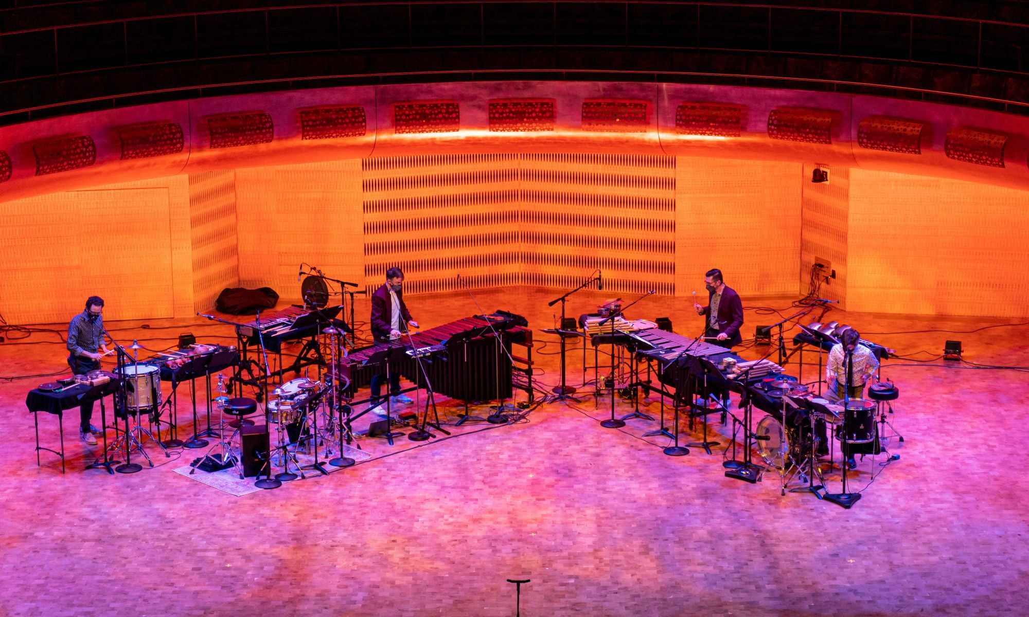 Four musicians playing a mix of percussion instruments under orange lighting.