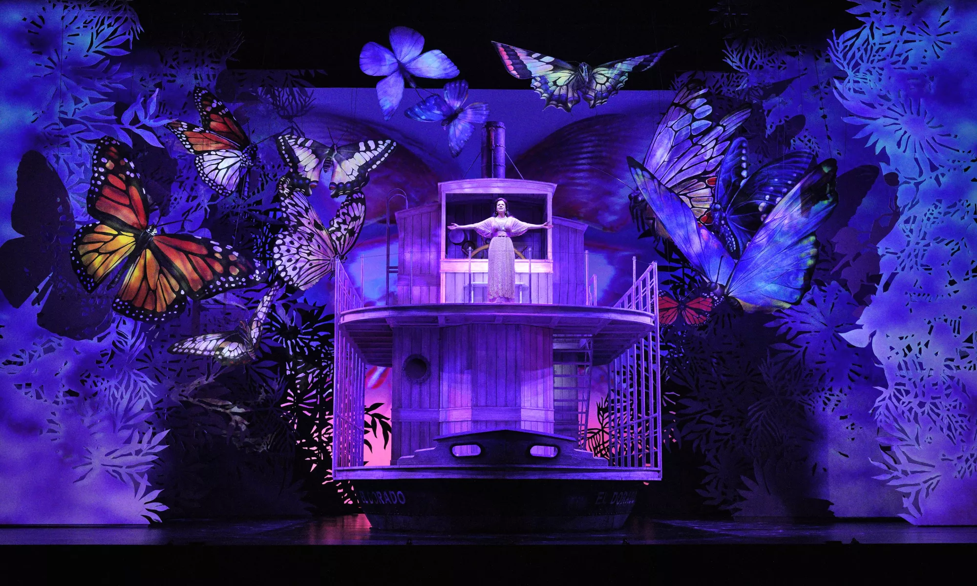 An image from the opera Florencia en el Amazonas: A woman stands with open arms at the front of a ship. She is surrounded by giant butterflies and illuminated in a purple light. 
