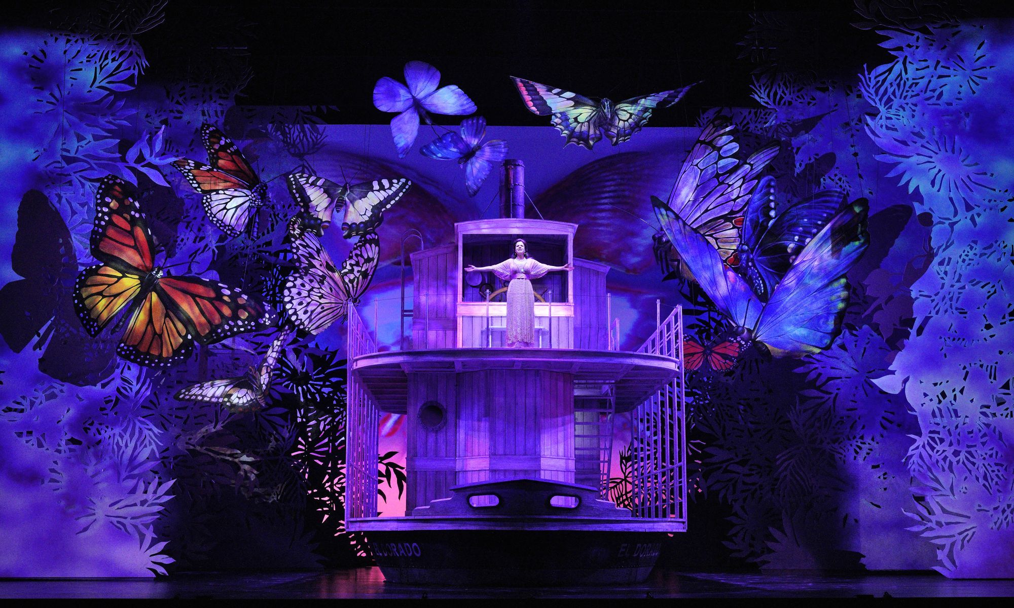 An image from the opera Florencia en el Amazonas: A woman stands with open arms at the front of a ship. She is surrounded by giant butterflies and illuminated in a purple light. 