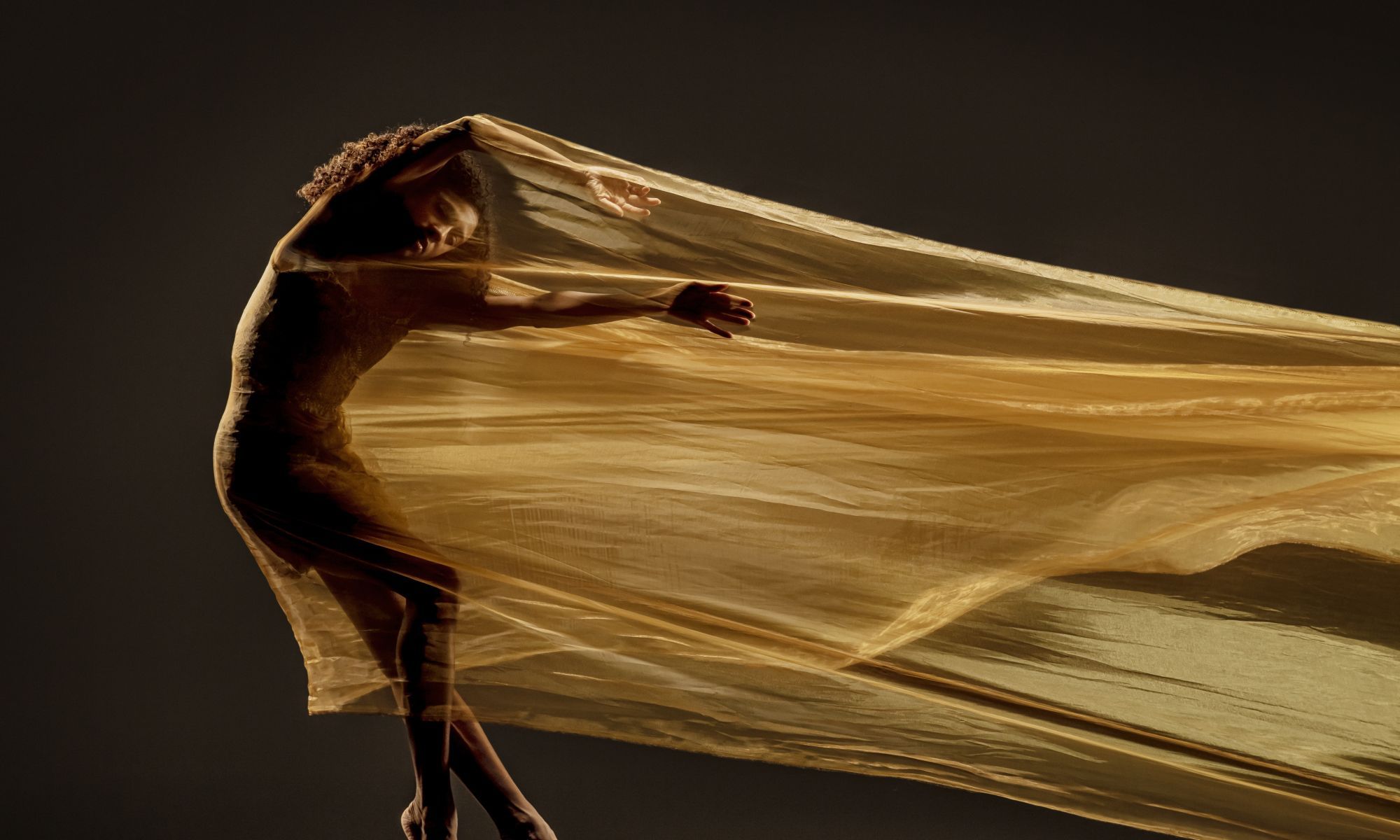 A Black dancer draped in gold fabric poses with outstretched arms