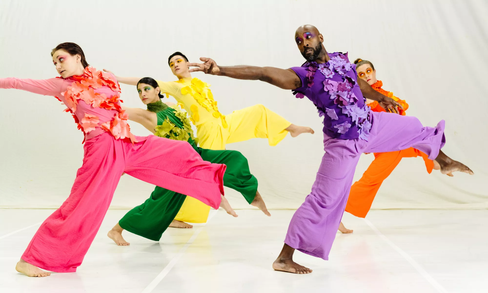 Five dancers in brightly colored, floral-appliqué costumes reach back with their arm while lunging forward with their leg. 
