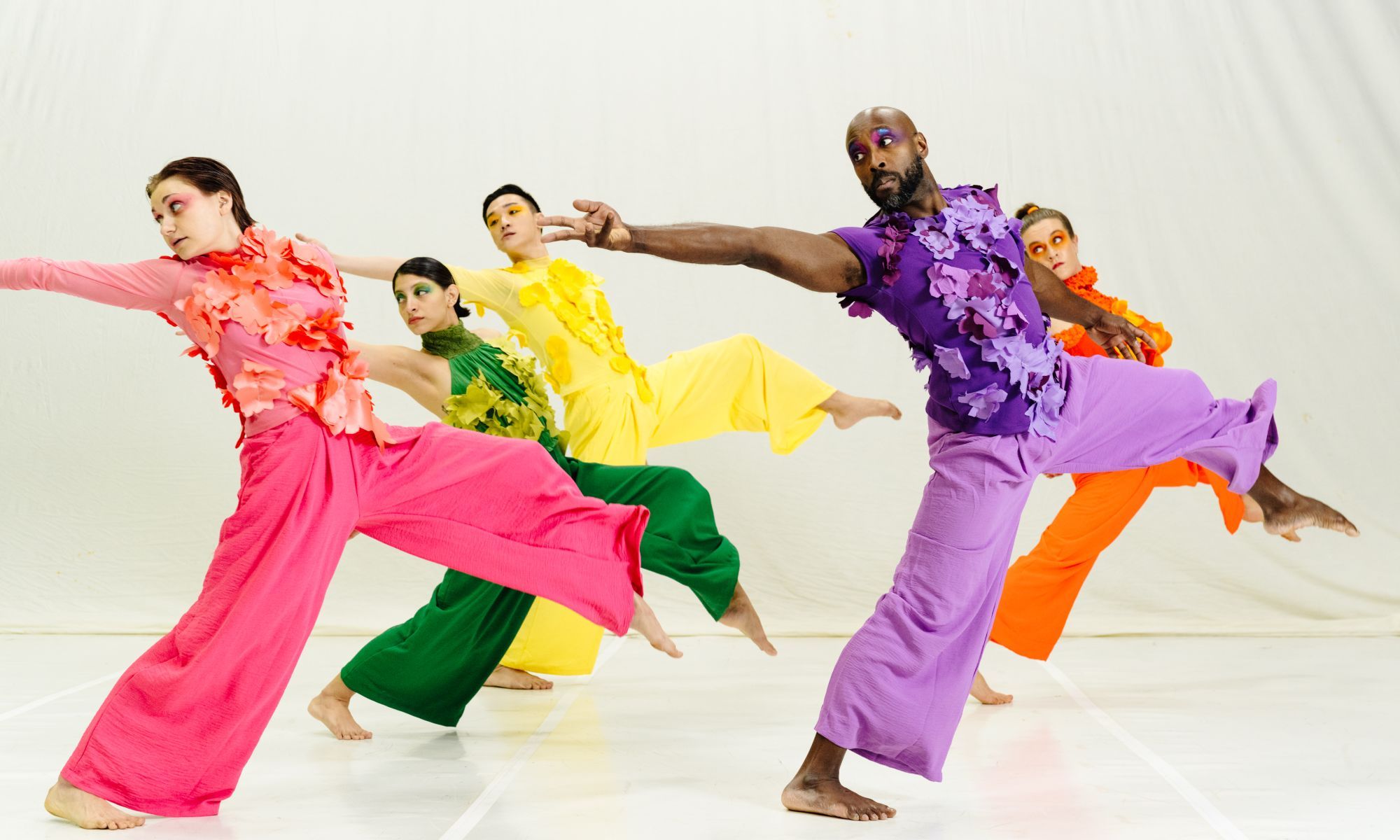 Five dancers in brightly colored, floral-appliqué costumes reach back with their arm while lunging forward with their leg. 