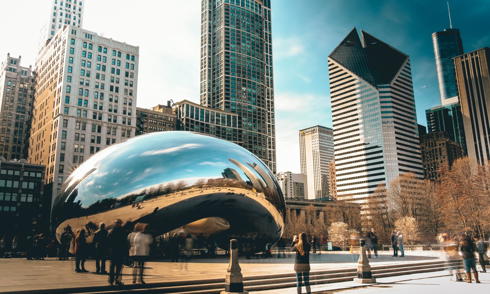 Image of Chicago's Cloudgate (The Bean) facing from the northwest. People are visible, taking pictures of the structure, and buildings can be seen in the background. 