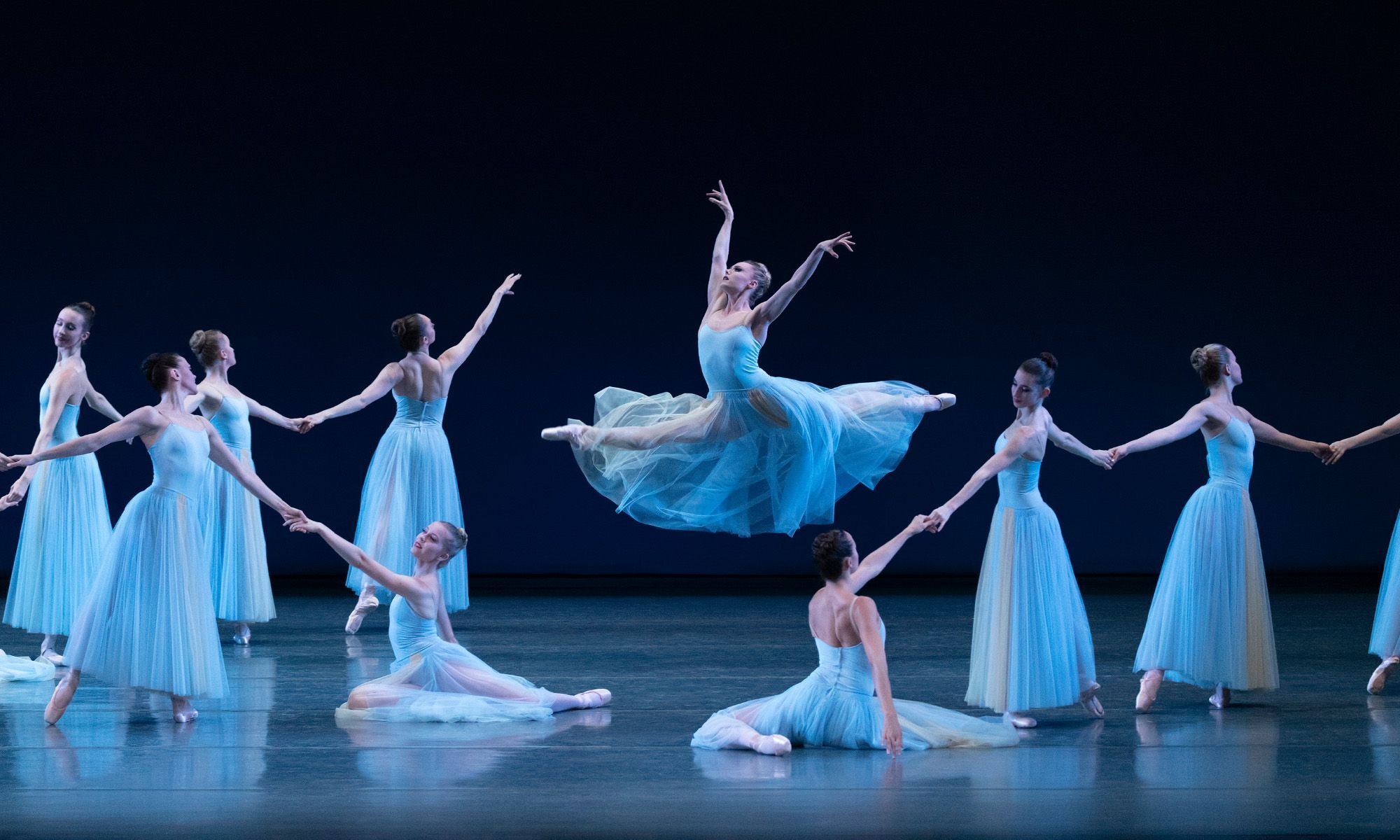 A group of ballerinas perform on stage holding hands as they circle one dancer. The dancer in the middle is mid-jump.