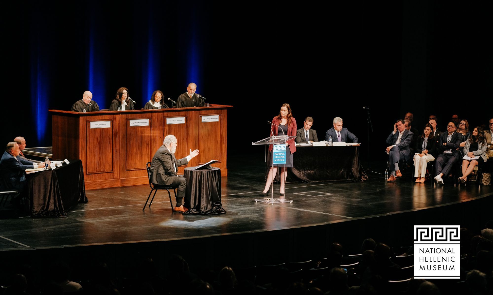 On a stage, a courtroom like setup takes place. 