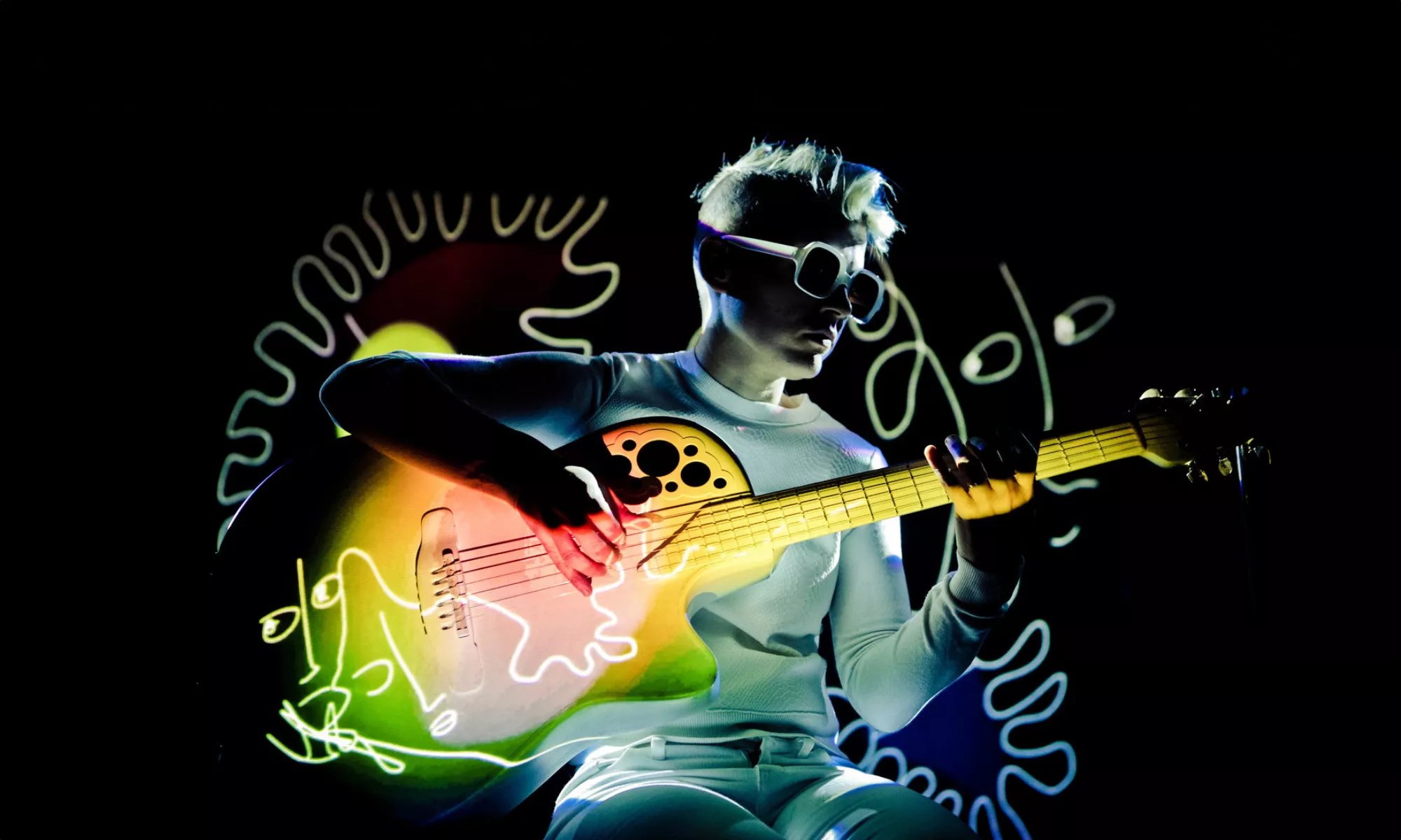 A person plays the guitar. A colorful illustration is projected on them and the guitar.