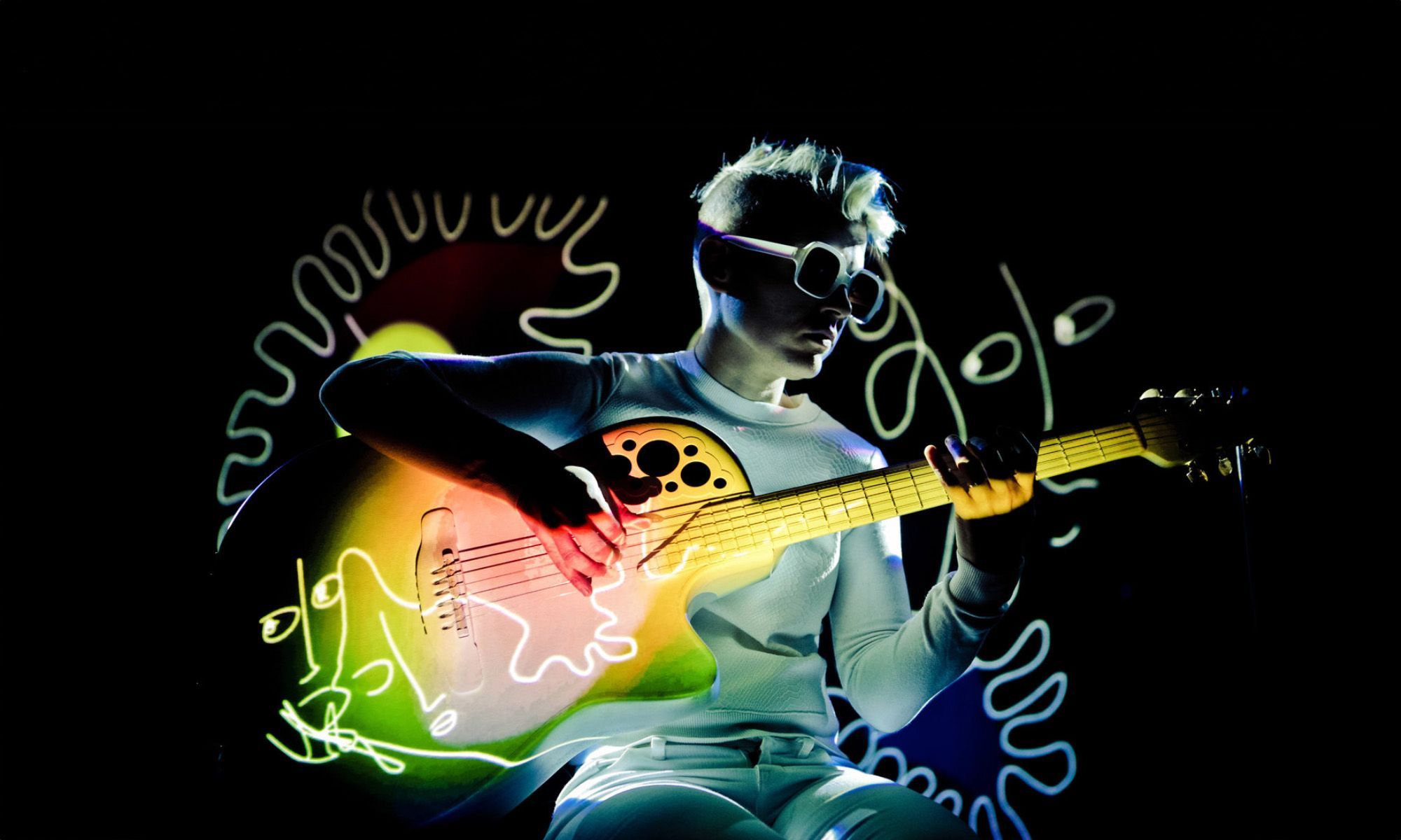 A person plays the guitar. A colorful illustration is projected on them and the guitar. 