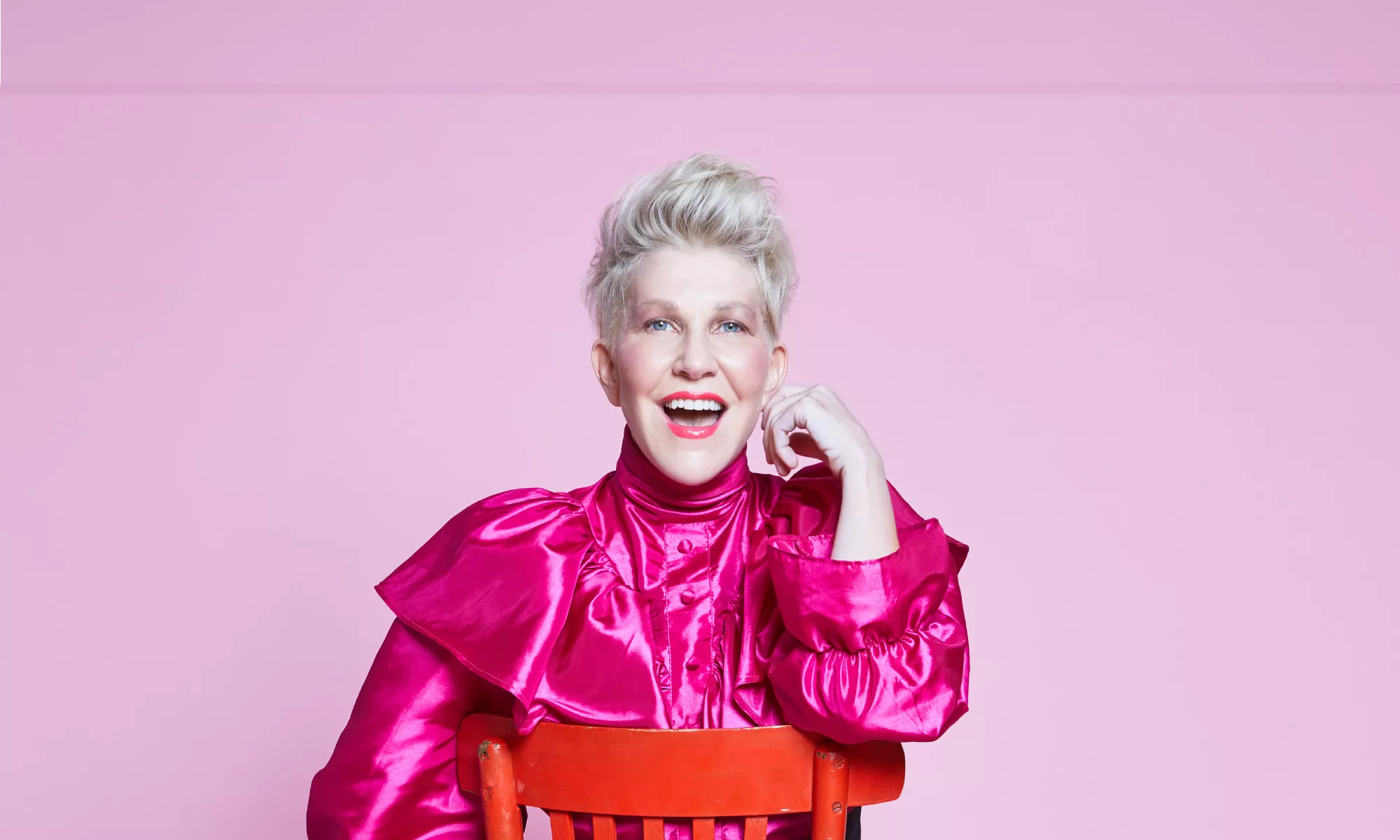 Joyce DiDonato smiling with her mouth open as she rests her hand on top of a blood orange chair.