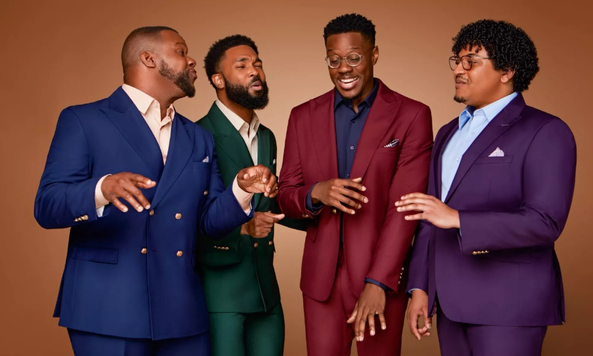 King's Return all wear different color suits. They stand in front of a solid brown backdrop as they sing.