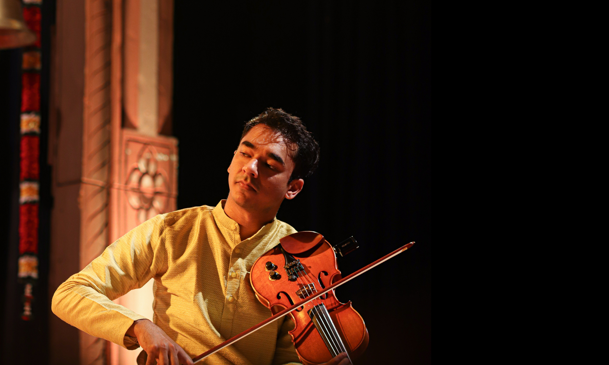 Ambi Subramaniam is sitting on stage playing violin in a yellow tunic. 