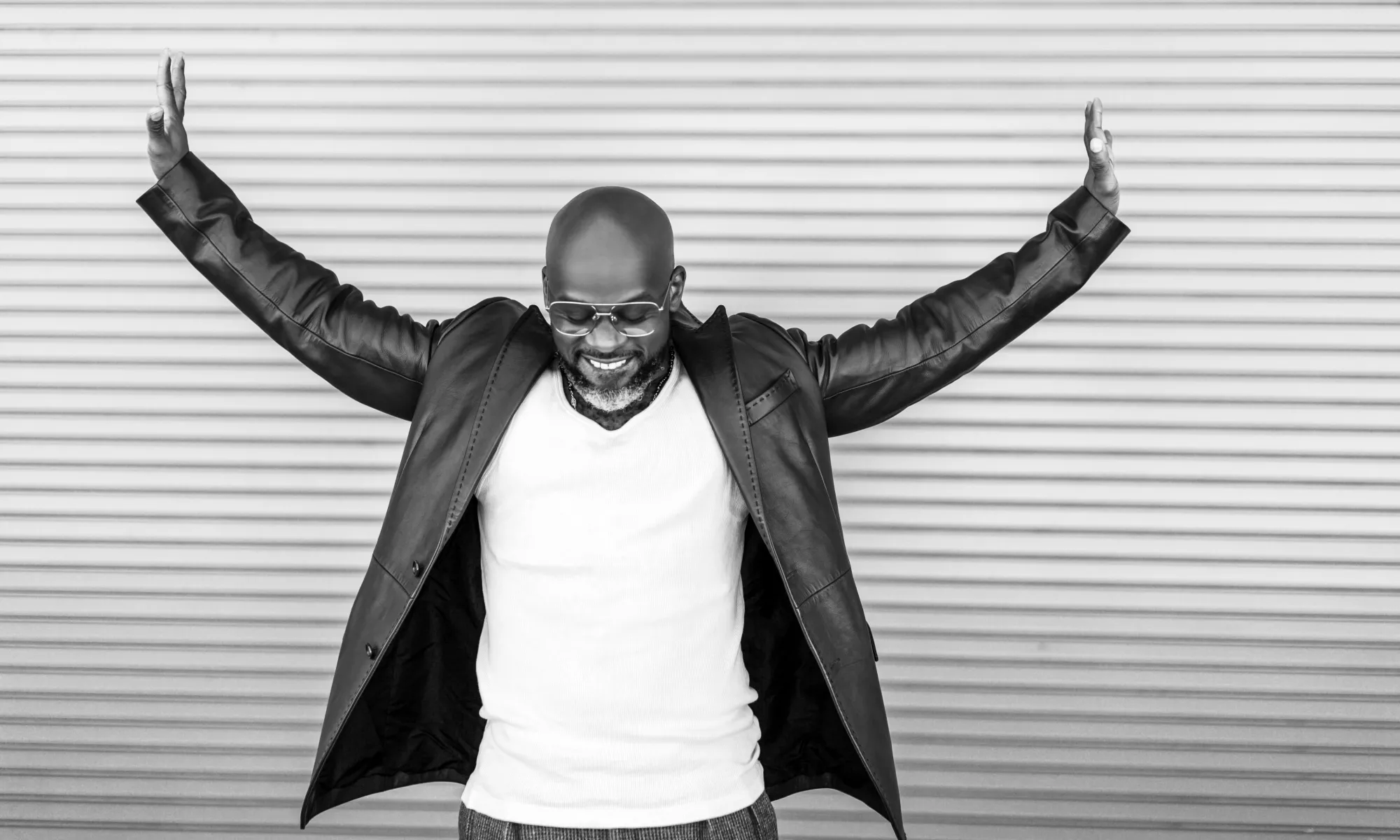 A black and white image of Marc Bamuthi Joseph while he lowers his face and extends his arms outward.